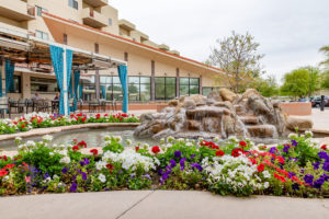 The exterior of the Friendship Village senior living center including red, white, and purple flowers and a natural rock landscape fountain