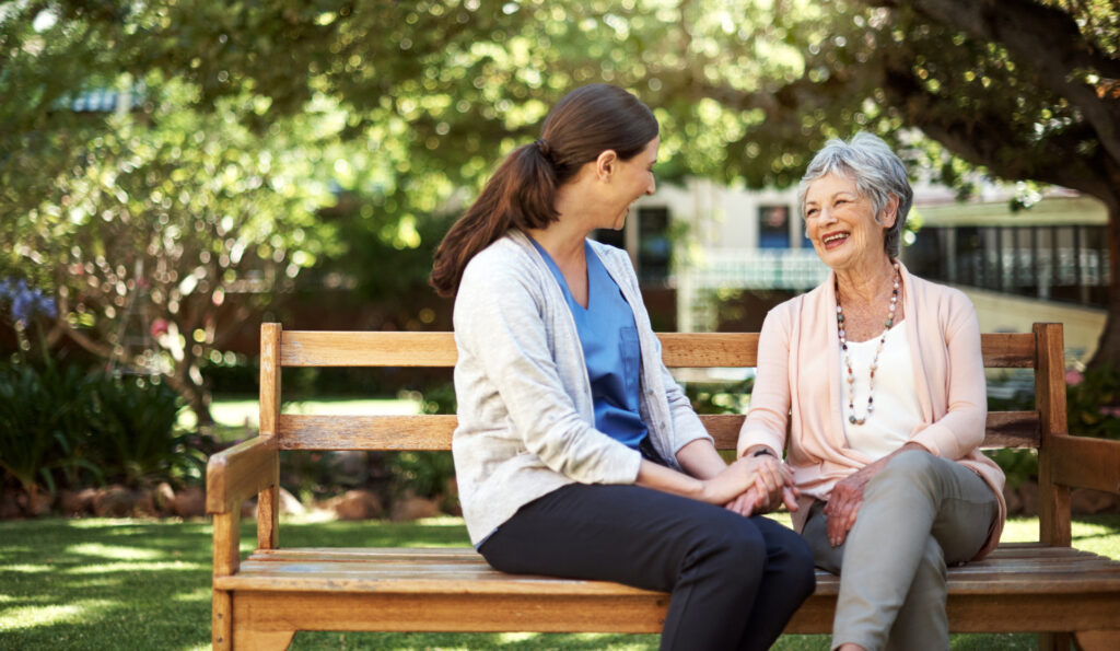 A health care worker and a senior woman sit on a park bench together