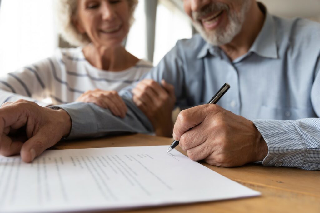 A senior man signs a contract while a senior woman sits next to him