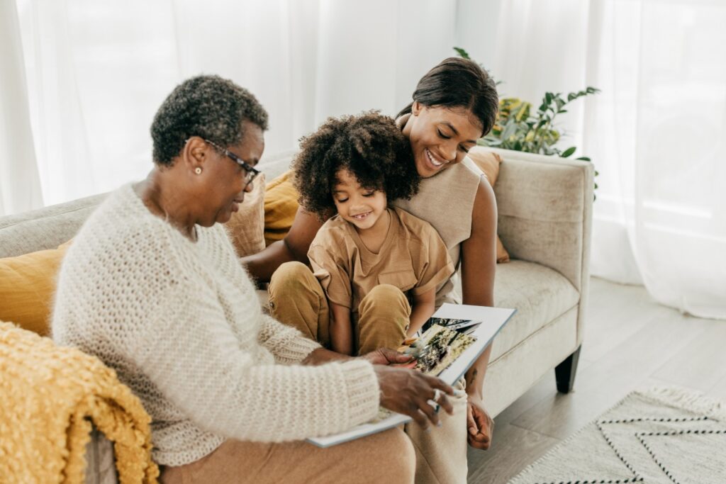 A woman, a grandchild, and a grandmother sit on couch and look at photos together