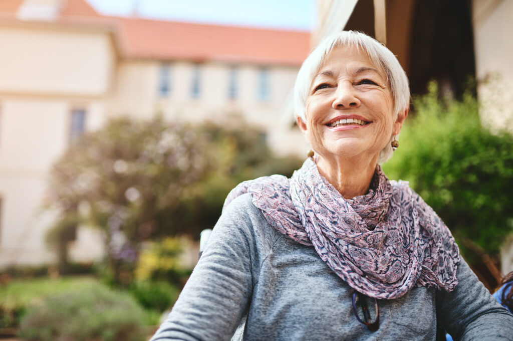 An older woman smiling while sitting outside of a multi-story senior living building