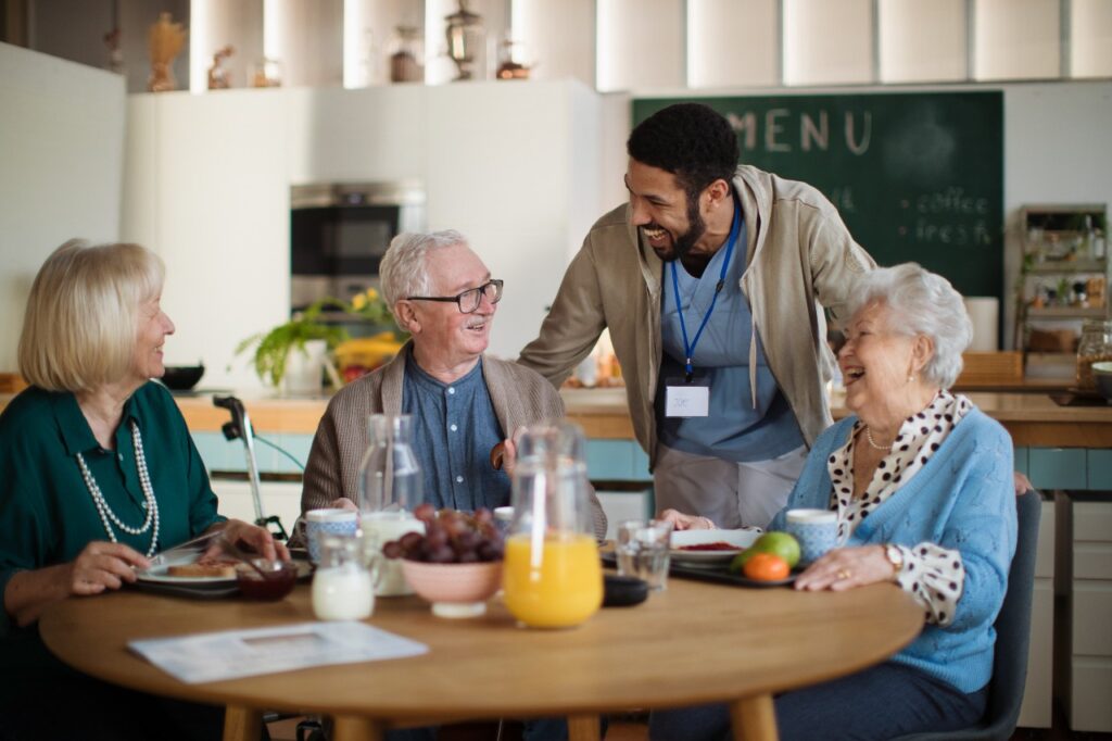 A group of seniors are enjoying a breakfast together at the table when a health care workers stops by to say hello